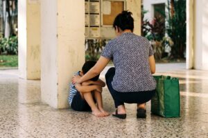 Mother crouching beside a young boy sitting on the floor of an HDB void deck in Singapore, comforting him as he buries his head in his arms